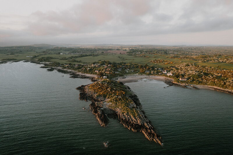 An aerial view shows a rocky, tree-covered headland jutting into the sea beside a coastal village and sandy beach.