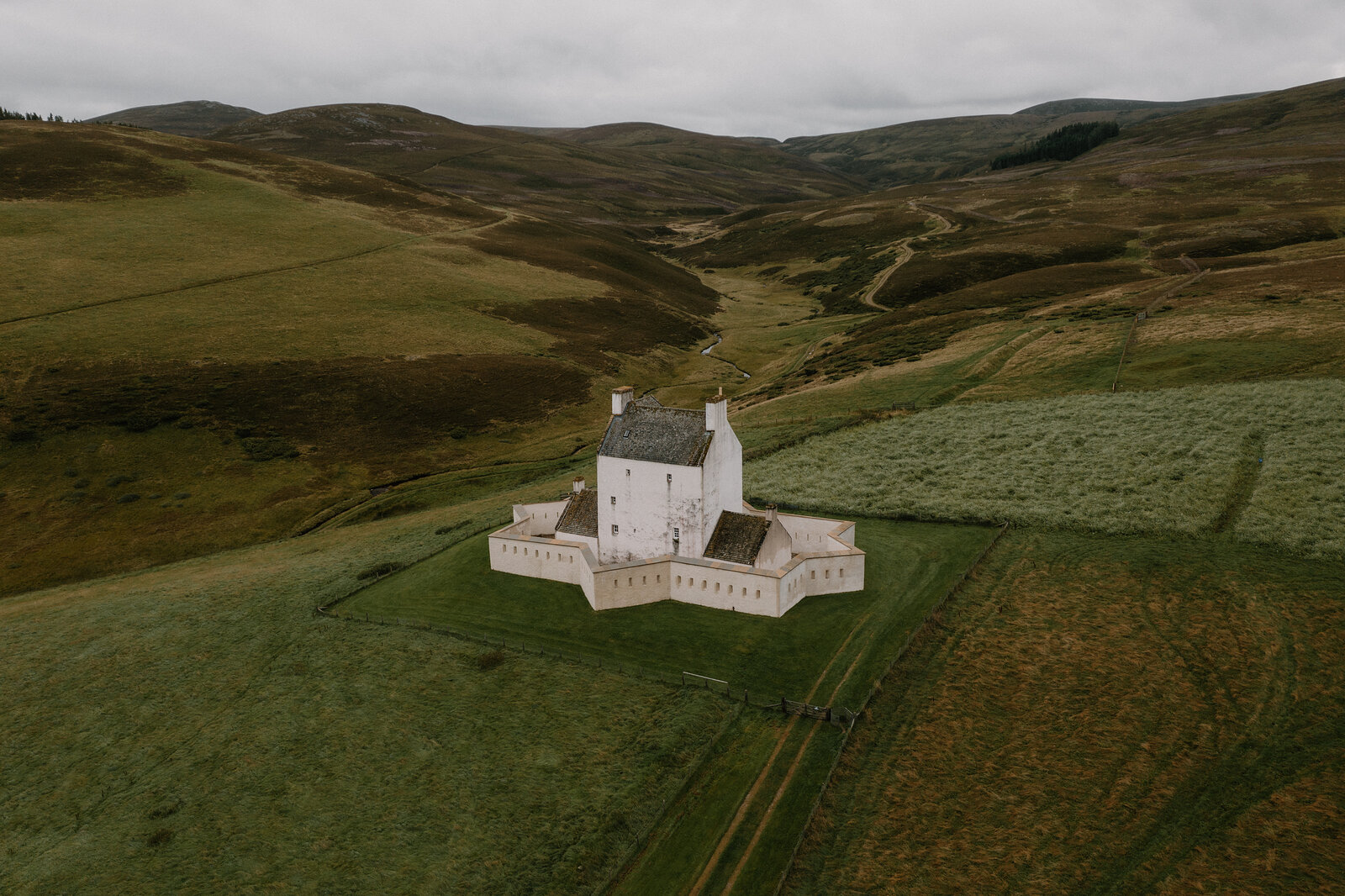A whitewashed star-shaped fortified tower house stands alone on a grassy hillside, hills rising behind under an overcast sky.
