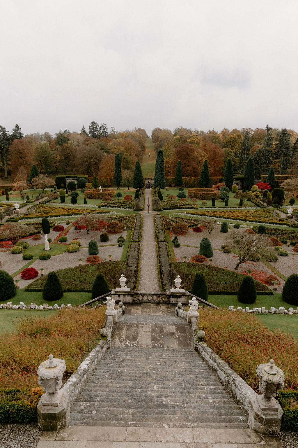 Stone steps flanked by urns and statues descend to a formal garden of clipped topiary and geometric flower beds, autumn trees framing the far edge.