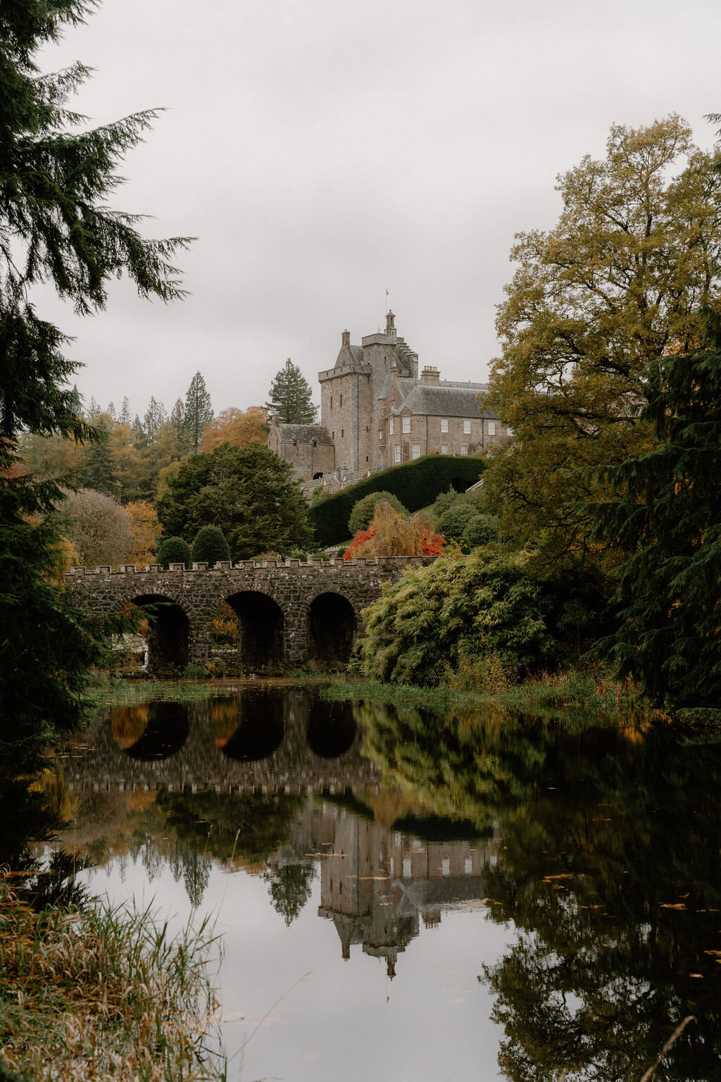 A castellated tower house with turrets stands on a wooded hillside beyond an arched stone bridge, autumn trees reflected in the still water below.