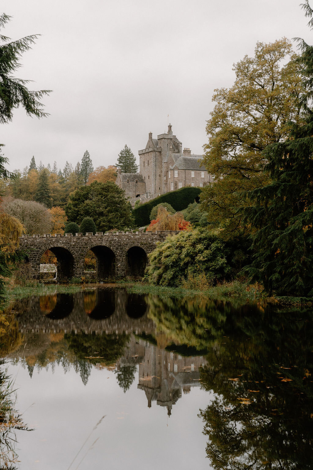 A castellated tower house stands on a wooded hillside beyond an arched stone bridge, autumn trees reflected in the still water below.