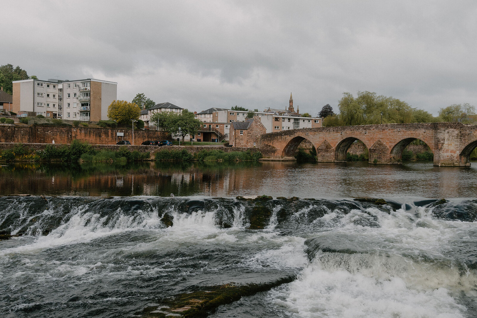 A river tumbles over a stepped weir toward an old stone multi-arched bridge, town buildings and a church spire lining the far bank.
