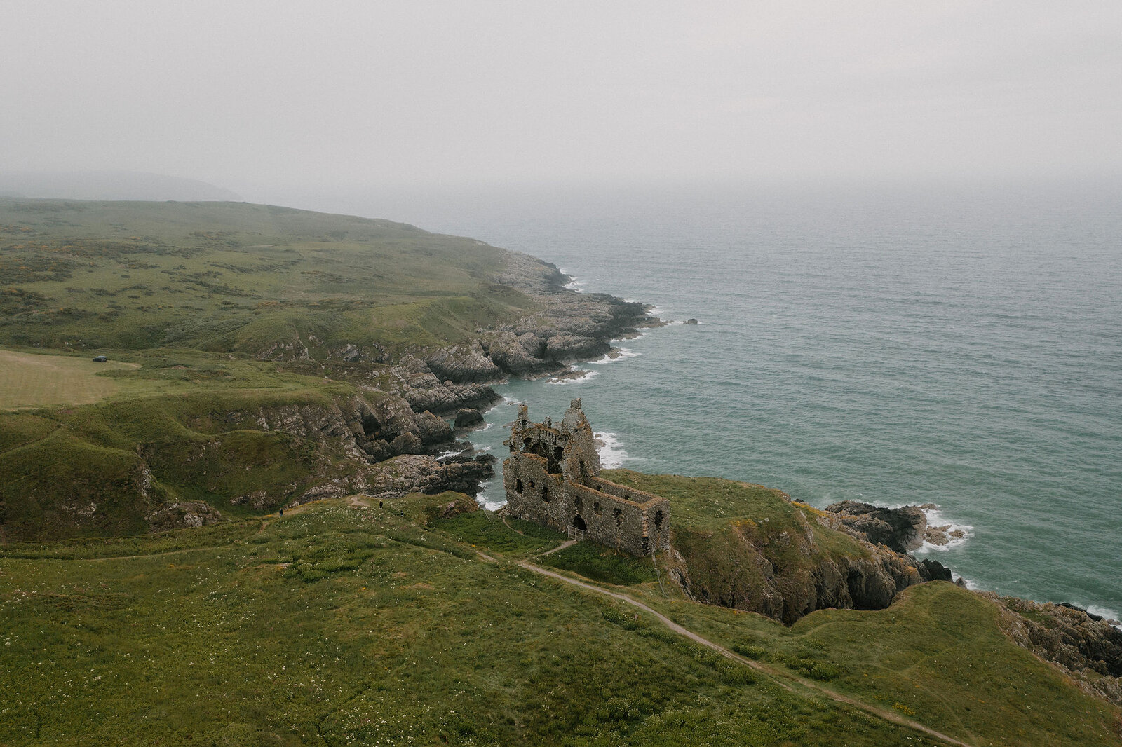 An aerial view in mist shows the ruined walls of a castle on a rocky headland, waves breaking along the cliffs and a path winding up from the shore.