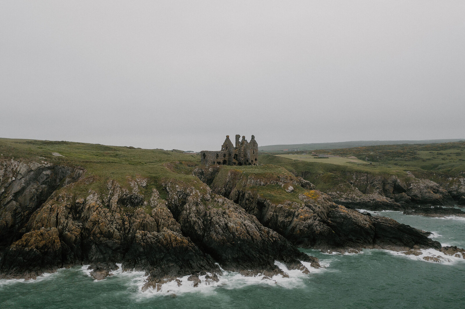 An aerial view in mist shows the ruined walls of a castle on a rocky headland, waves breaking along the cliffs below.