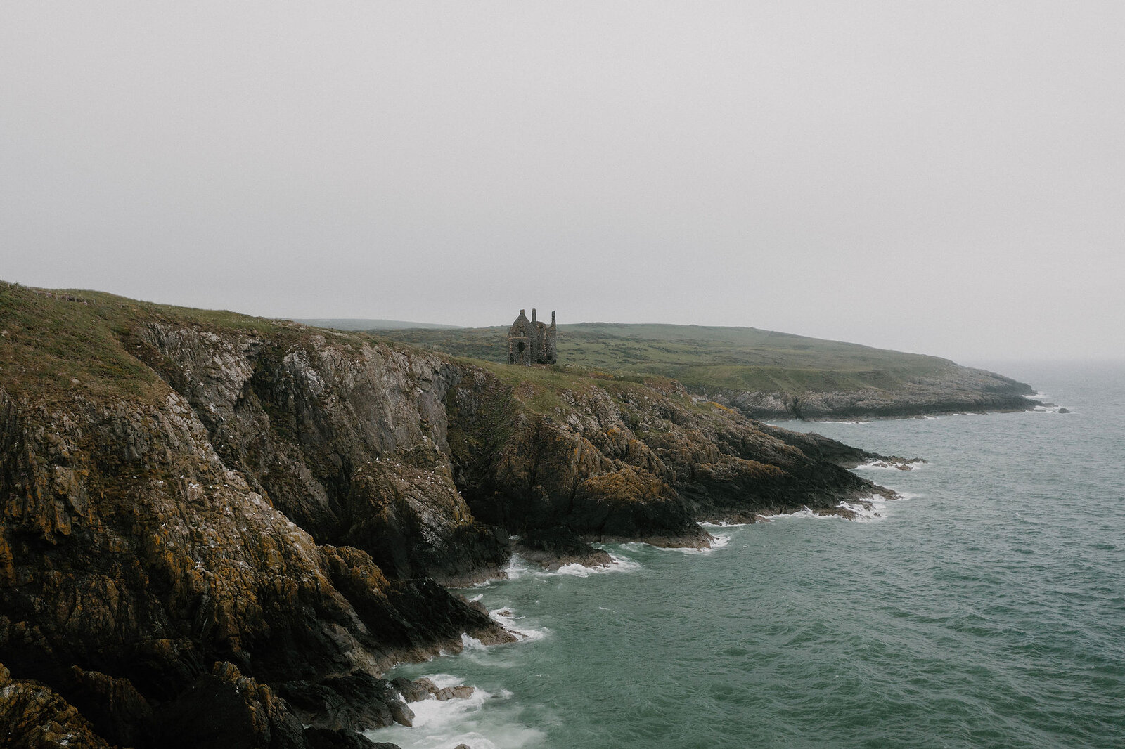 The ruined walls of a small chapel stand at the edge of a grassy cliff above the sea, mist softening the coastline behind.