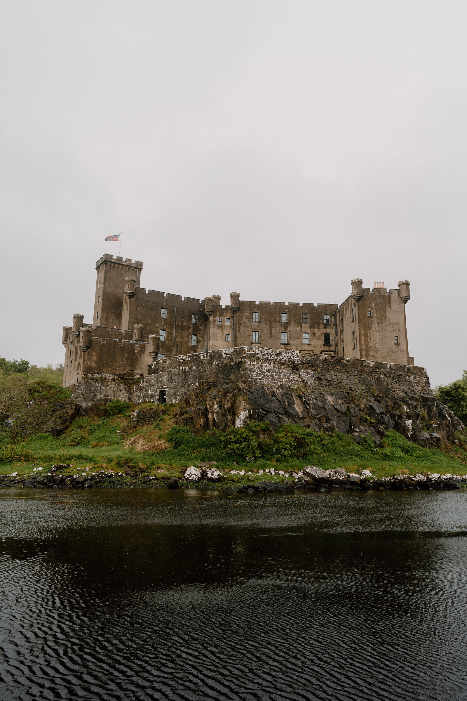 A castle with battlements and turrets stands on a rocky outcrop rising from calm water, a flag flying above.