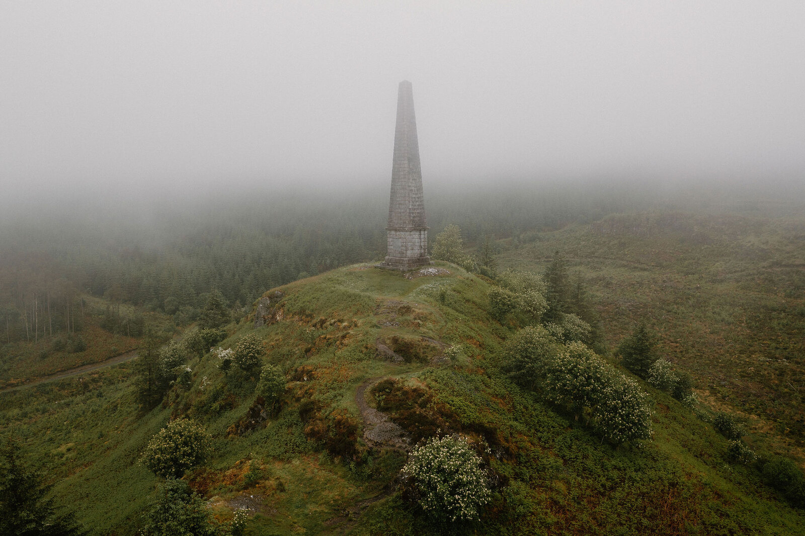 An aerial view shows a stone obelisk monument standing on a misty hilltop, forest and hawthorn blossom surrounding its base.