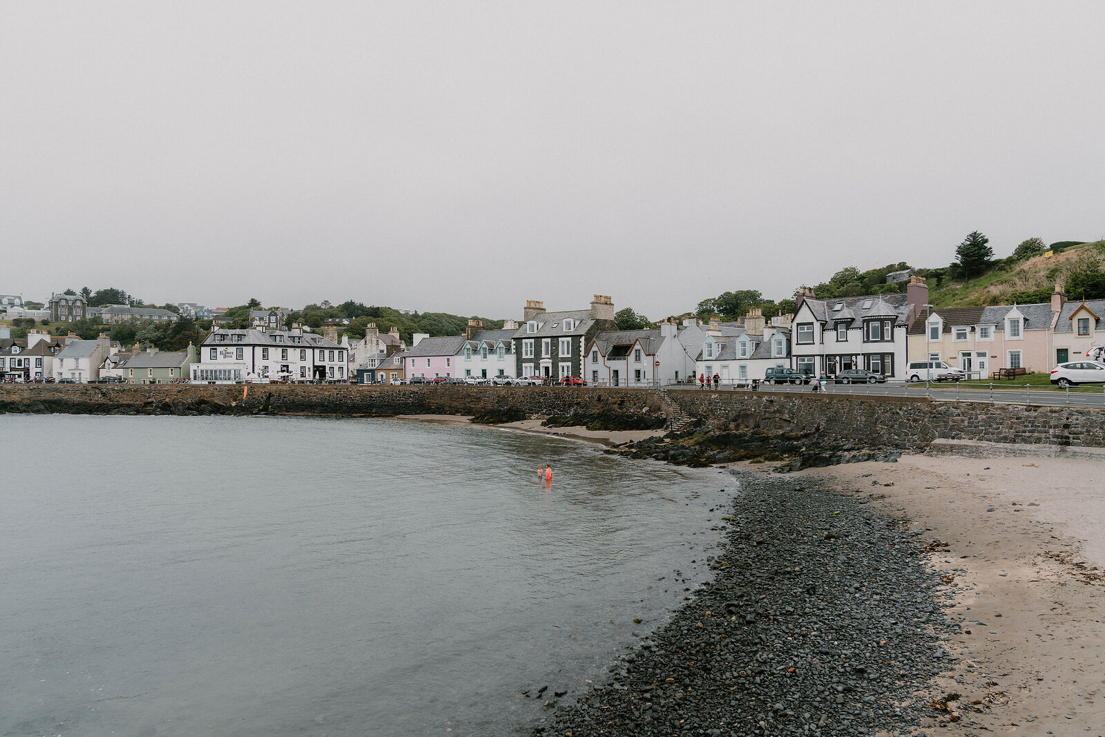 A harbour village of colourful houses lines a shingle beach, wooded hills rising behind under an overcast sky.