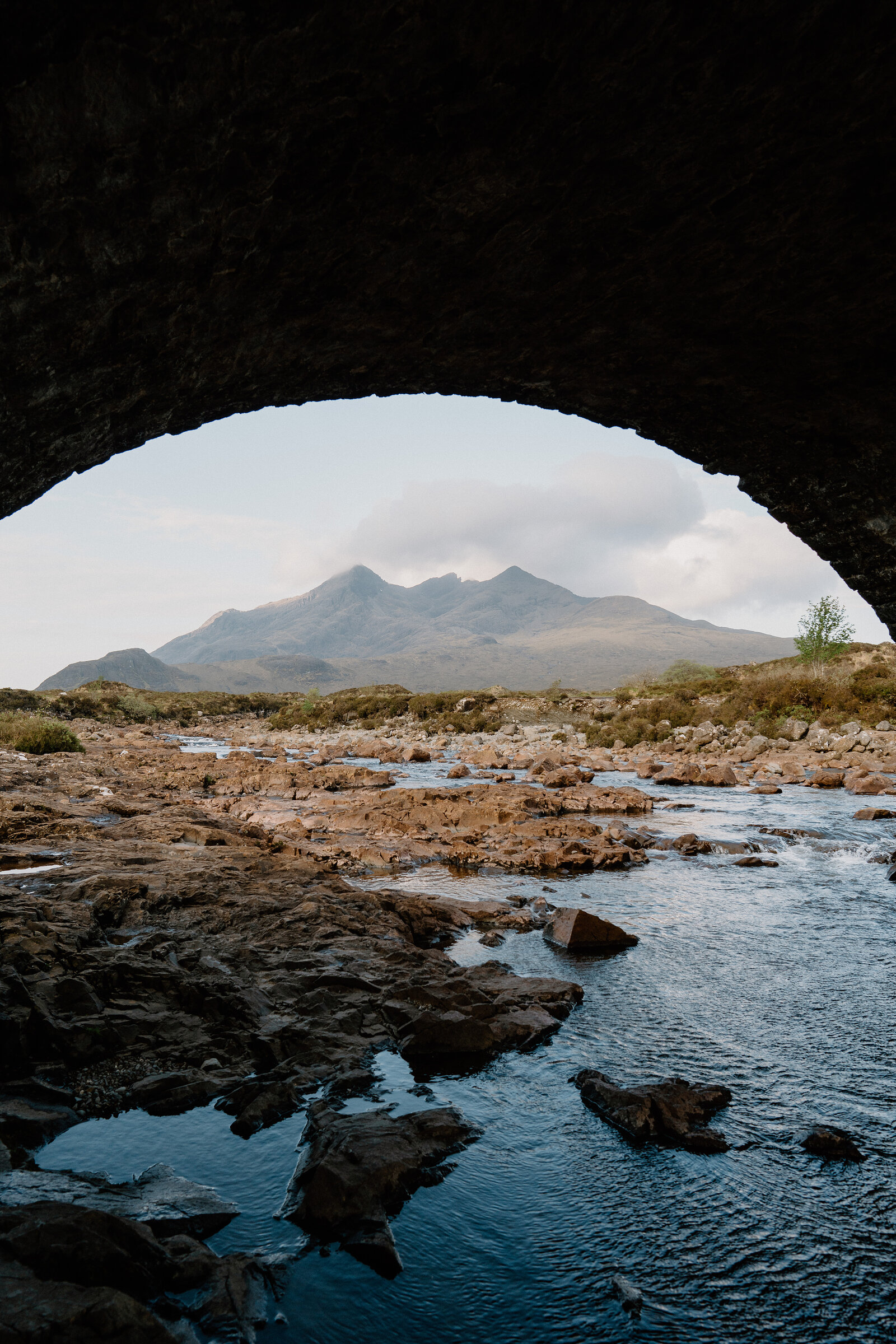 The stone arch of a bridge frames a view of jagged mountain peaks beyond a rocky, shallow river.