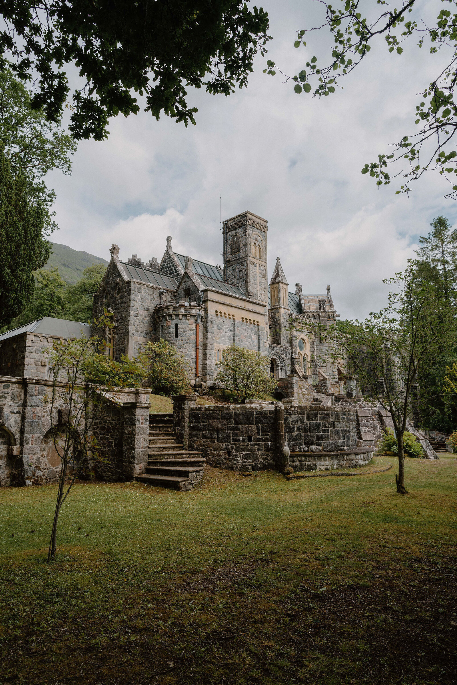 A stone church with a tall tower and buttressed cloister stands among trees, stone steps leading up to it from a lawn.