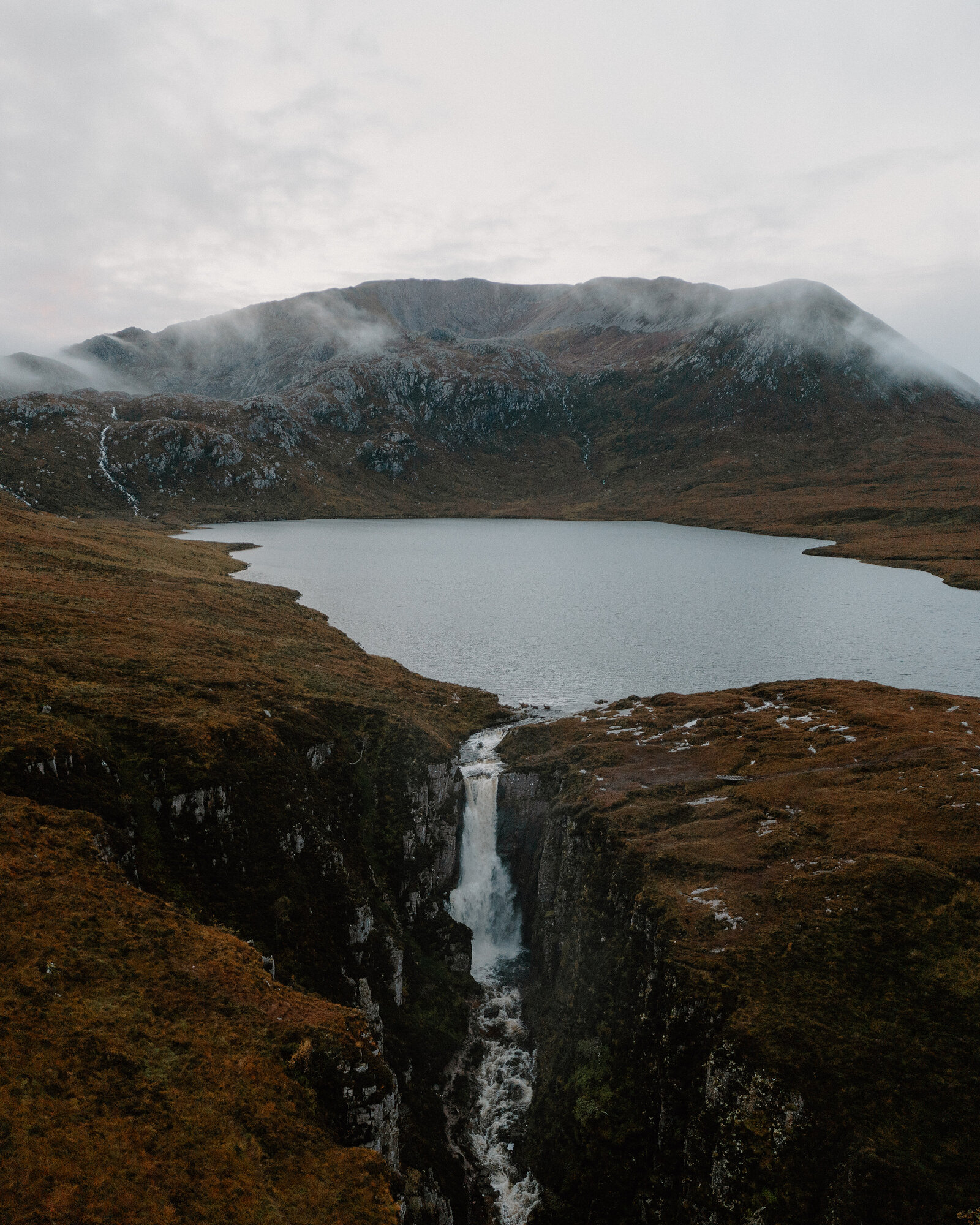 A waterfall spills from a loch over a rocky edge into a gorge below, a misty mountain rising behind moorland streaked with heather.