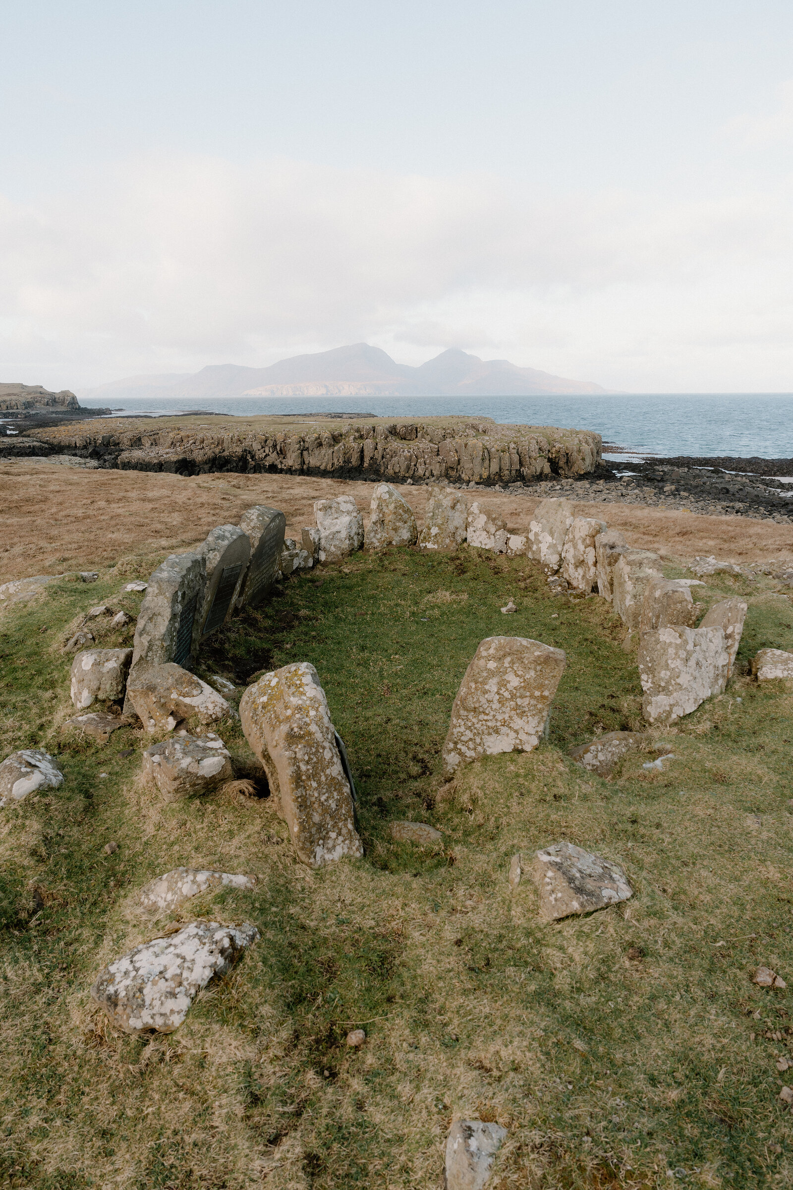 The circular stone foundation of an ancient roundhouse sits on a coastal headland, distant mountains visible across the sea.