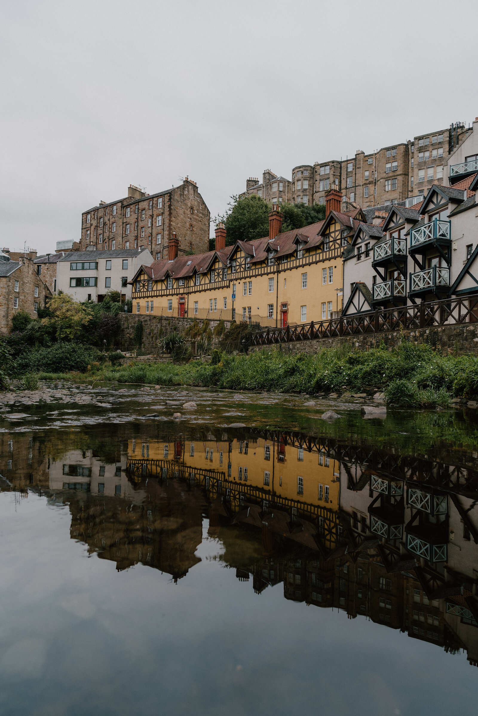 Colourful half-timbered buildings with wrought-iron balconies line a river, tenement buildings rising on the hillside behind and reflecting in the water.