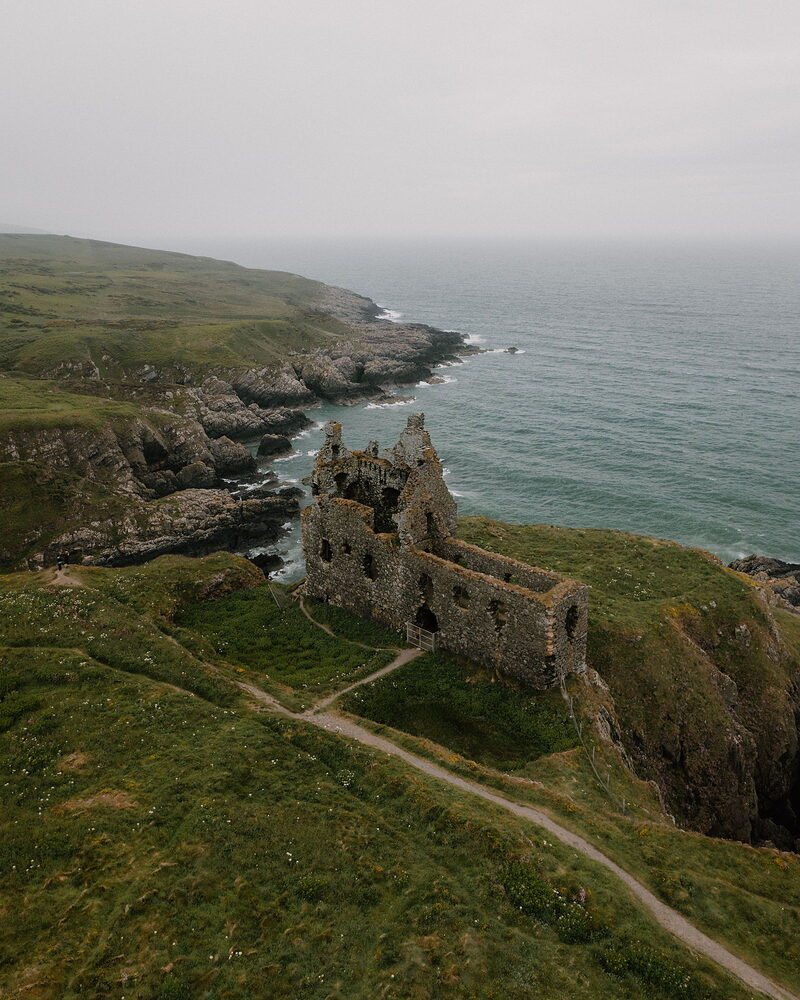 An aerial view shows a ruined castle standing on a grassy coastal headland, cliffs dropping to the sea on either side.
