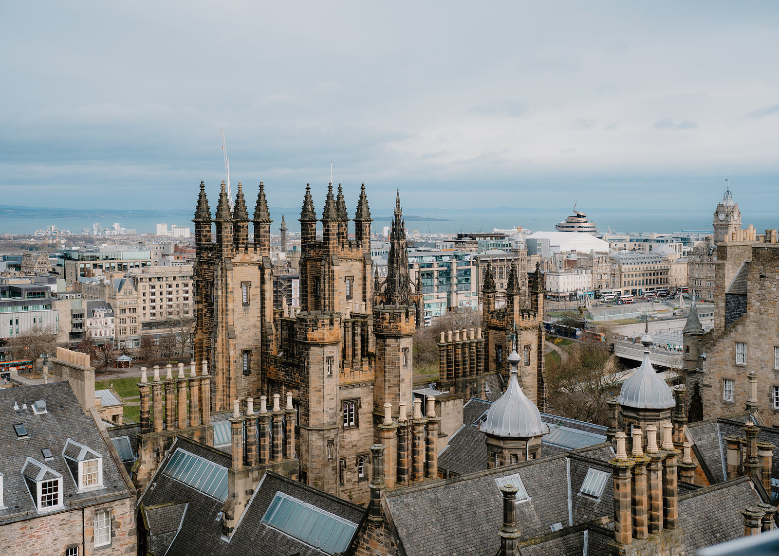 Gothic spires and pinnacled towers rise above a rooftop of skylights, the city and firth spreading beyond.