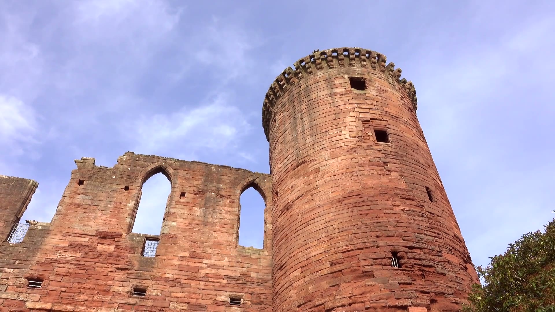 Looking up at a round tower of red sandstone at Bothwell Castle and the tall pointed windows of the wall beside it, clouds crossing the sky.