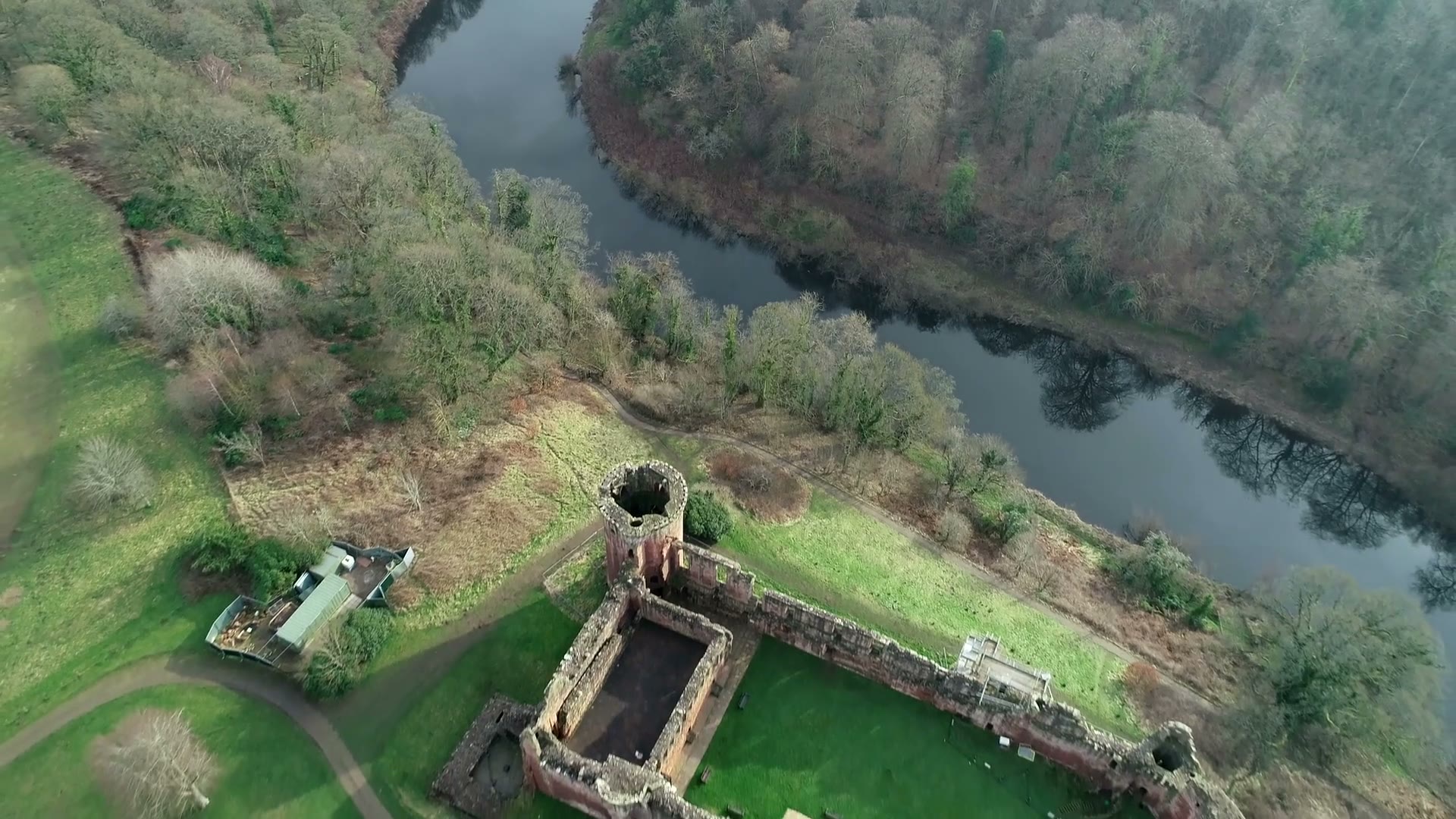 Bothwell Castle from high above, its walls and round towers round the green courtyard, the River Clyde curving through the woods beyond.