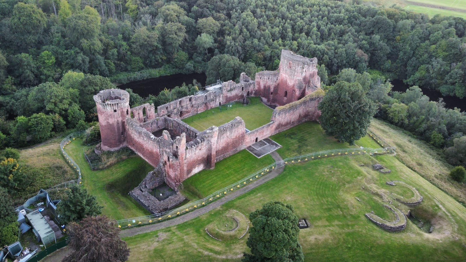 Bothwell Castle from the air, its red sandstone curtain walls and round towers round a green courtyard, the River Clyde dark among the woods behind.