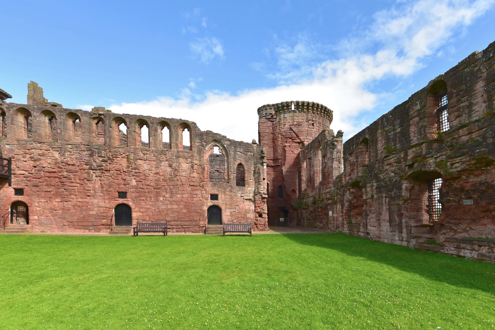 Inside the courtyard of Bothwell Castle on a summer day, a wall of tall arched windows and a round tower in red sandstone above the green lawn.