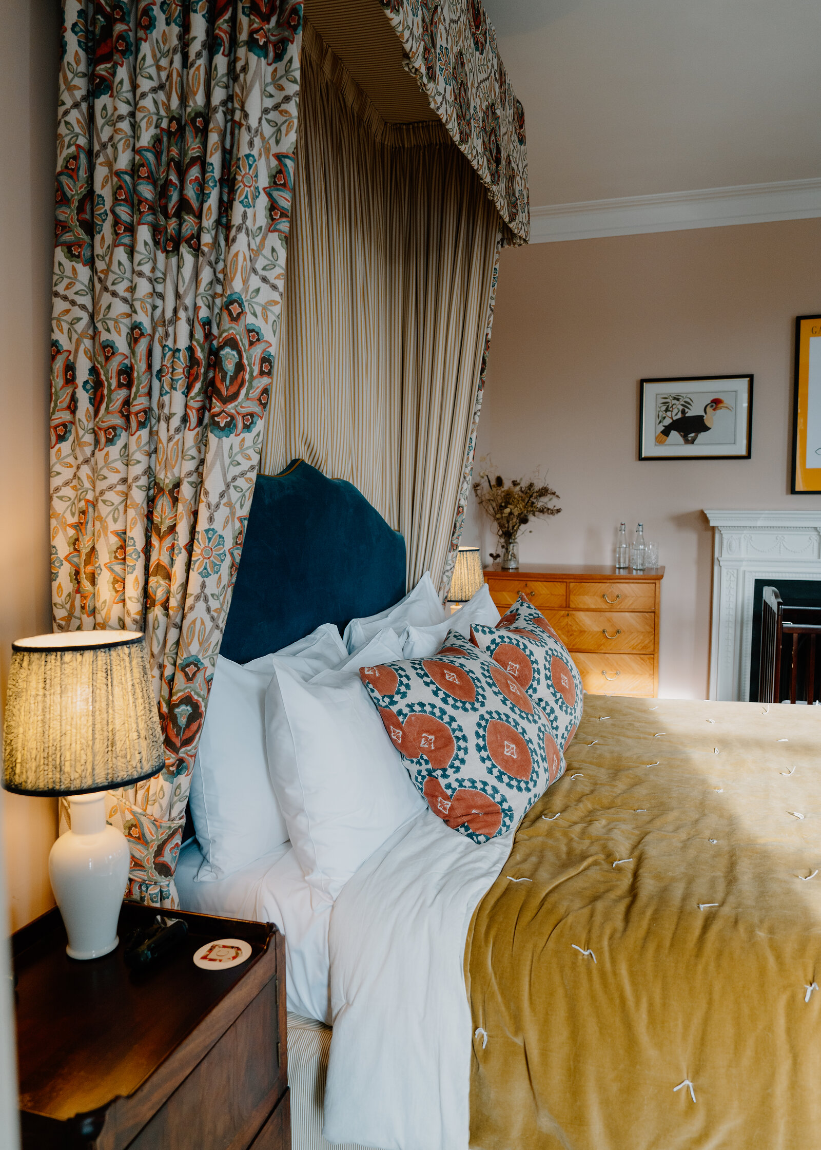 A four-poster bed at Cromlix dressed in white linen and a mustard velvet throw, beside floral canopy drapes and a lit bedside lamp