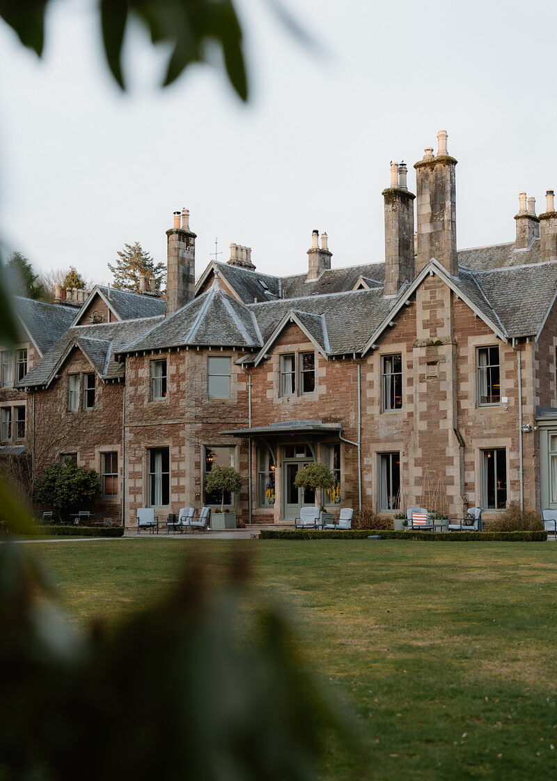 A turreted sandstone mansion with rows of tall chimneys rises behind a lawn, garden chairs set along its terrace.