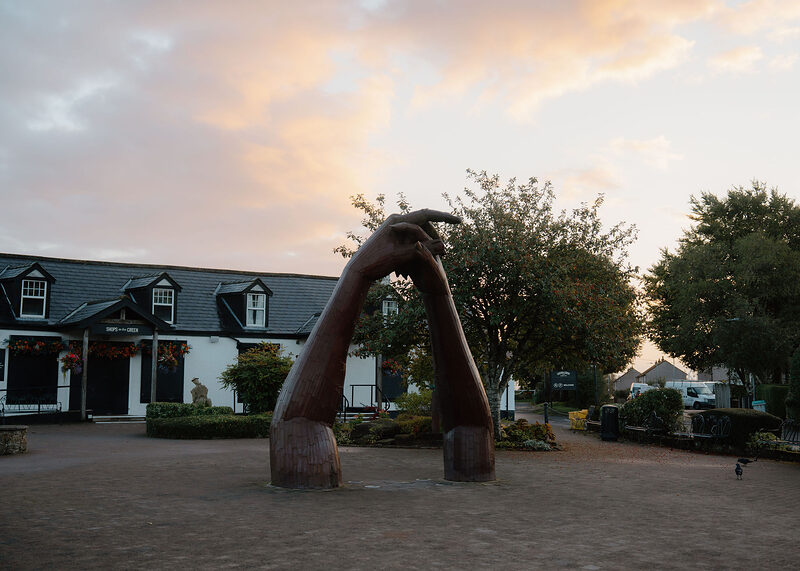 The Big Dance, the giant steel clasped hands, in the Gretna Green square at sunset, with the black-and-white shops on the left and a crow on the cobbles.