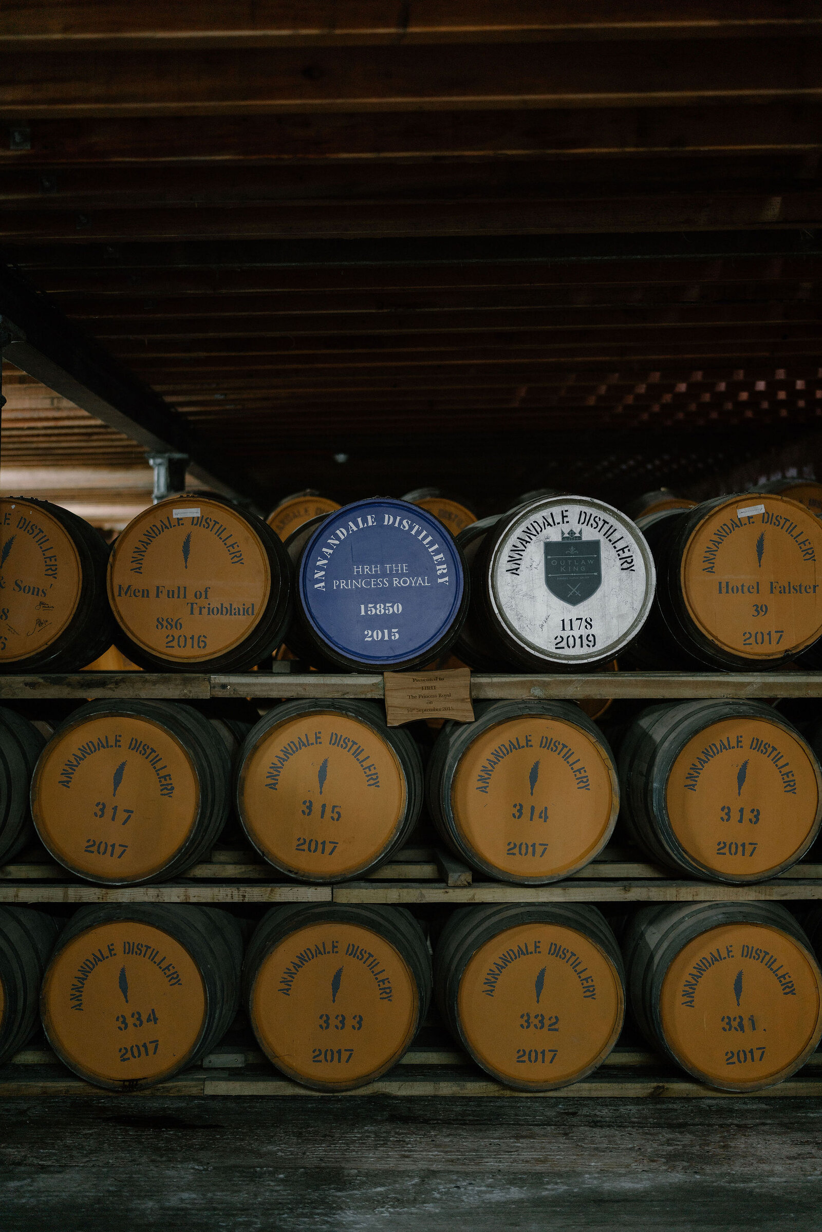 Named whisky casks including one marked HRH The Princess Royal stand stacked in a warehouse, branded plaques and dates identifying each one.