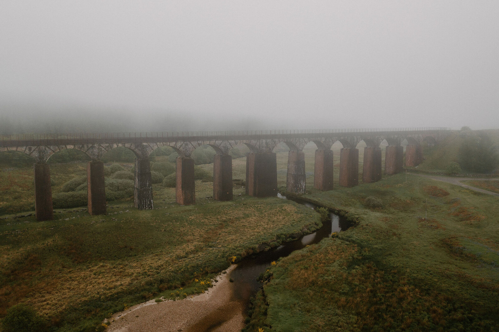 A tall brick and stone railway viaduct strides across an open field through thick mist, a stream winding beneath its arches.
