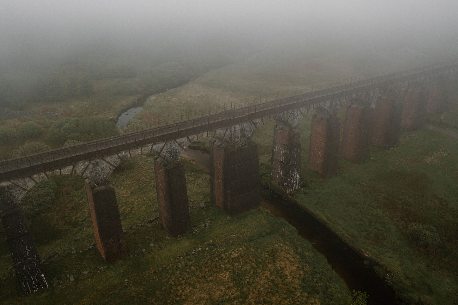 An aerial view shows a tall brick and stone railway viaduct striding through thick mist over a stream and grassy field.