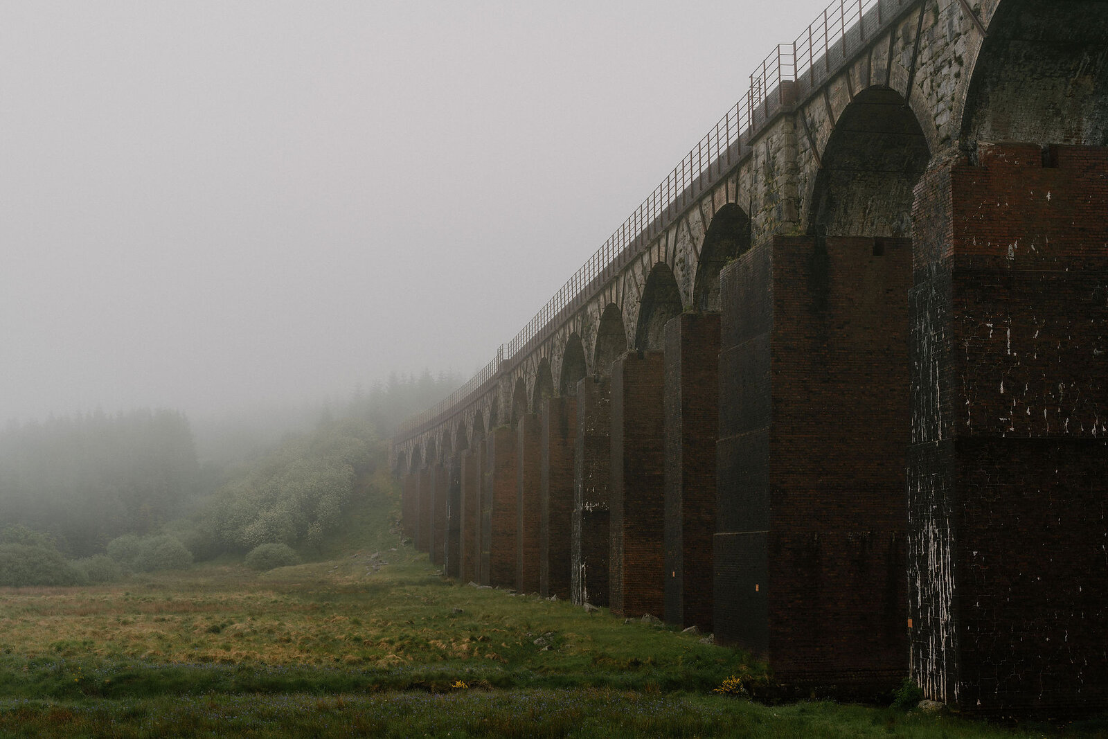 A tall brick and stone railway viaduct recedes into thick mist, its many arches striding across an open grassy field.