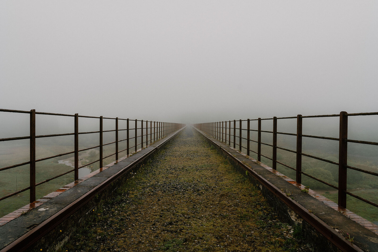 A footbridge with metal railings stretches into thick fog, its walkway vanishing where the mist closes in.