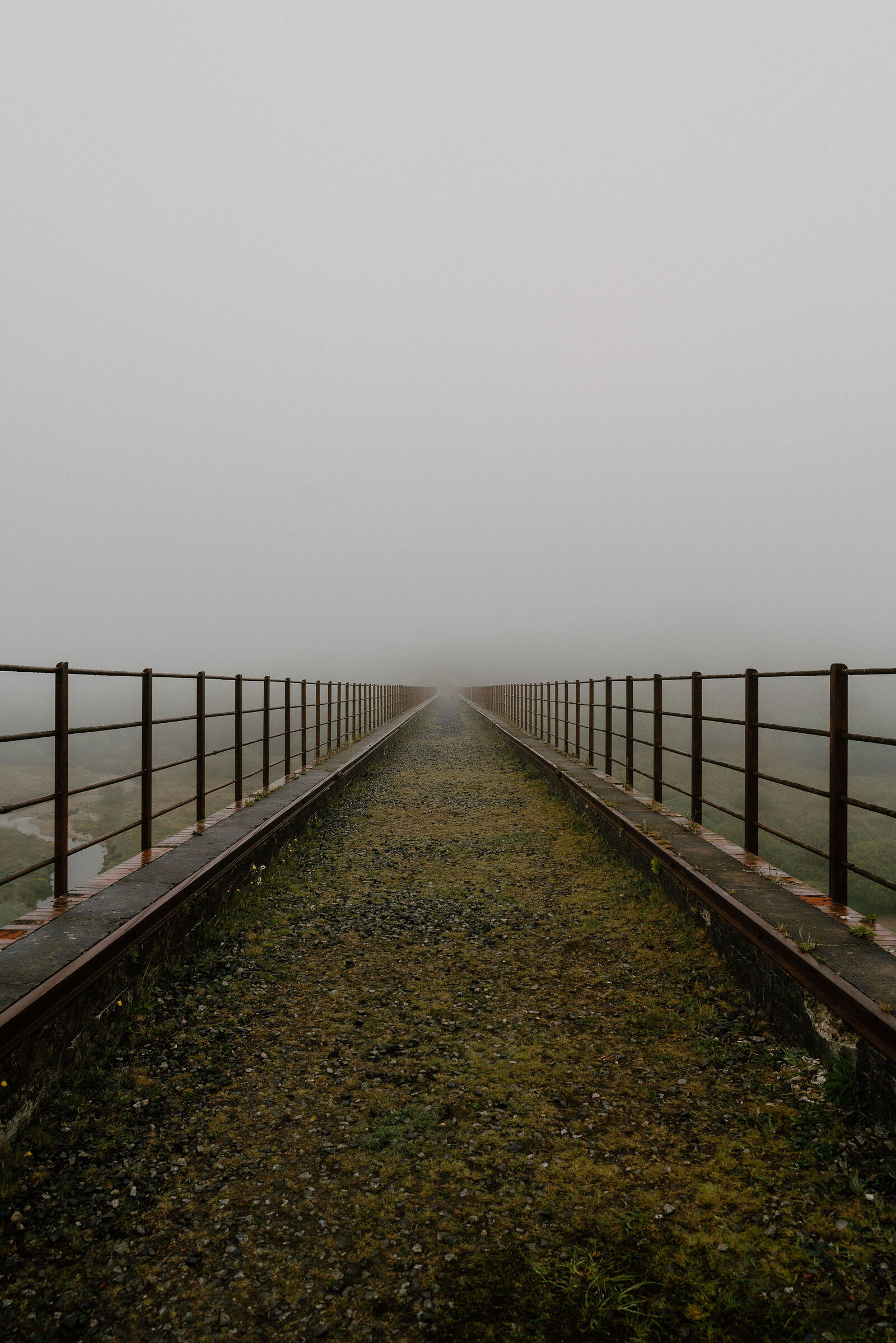 A footbridge with rusted metal railings stretches into thick fog, its stone walkway disappearing where the mist closes in.