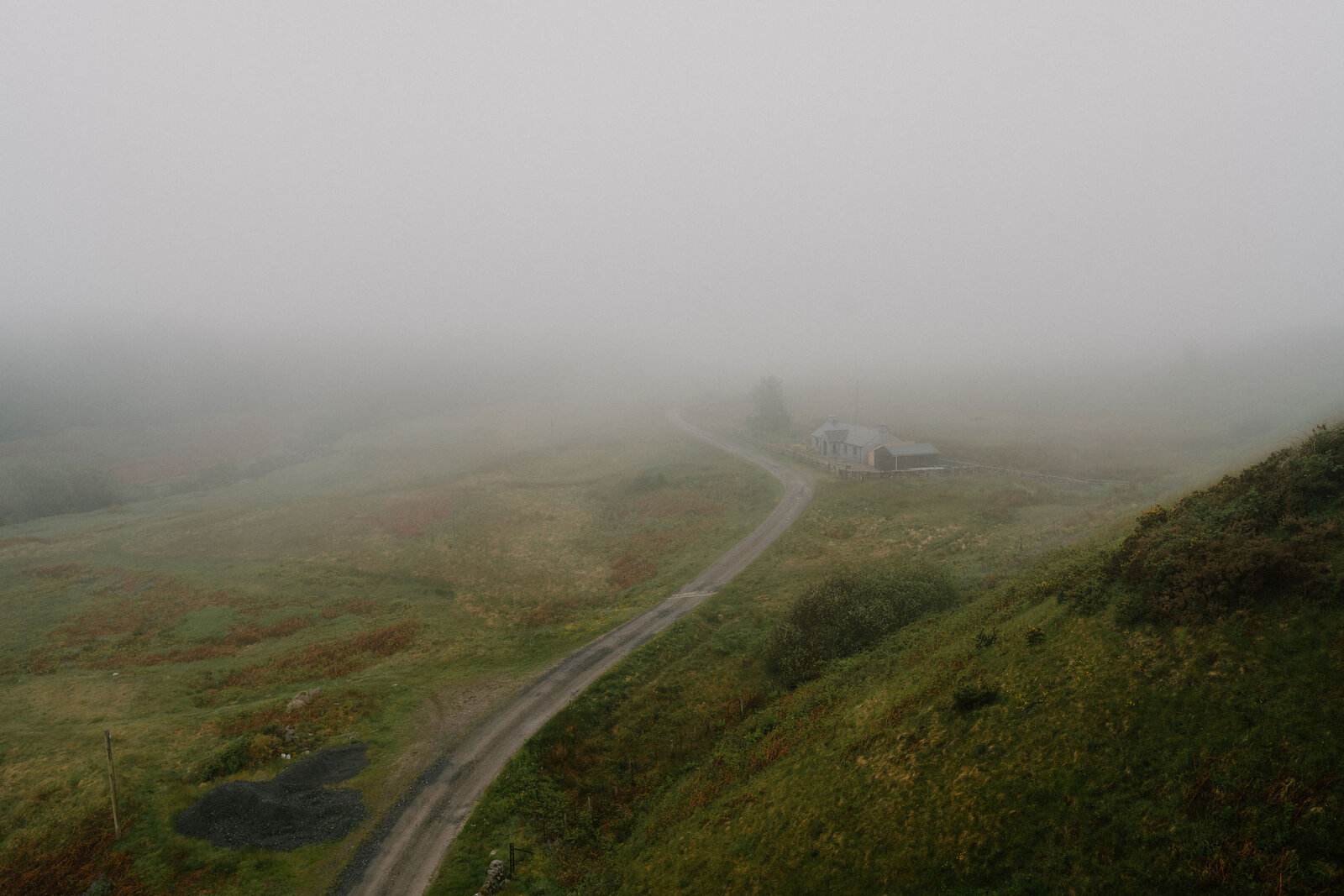 A road winds through misty moorland towards a building fading into the fog, heather covering the slopes on either side.