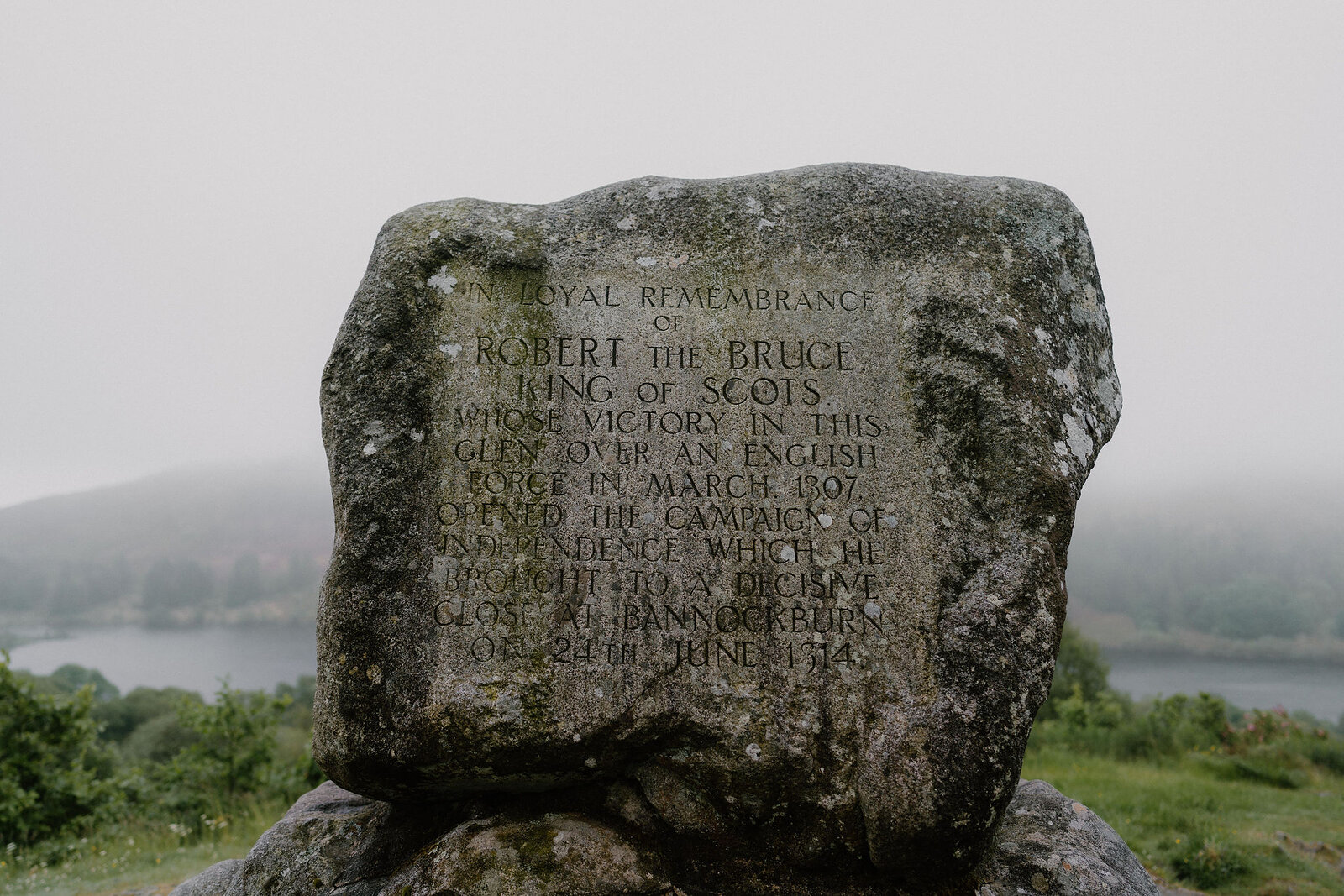 A weathered stone monument inscribed "In loyal remembrance of Robert the Bruce, King of Scots, whose victory in this glen over an English force in March 1307..." stands on a hillside above a loch in mist.