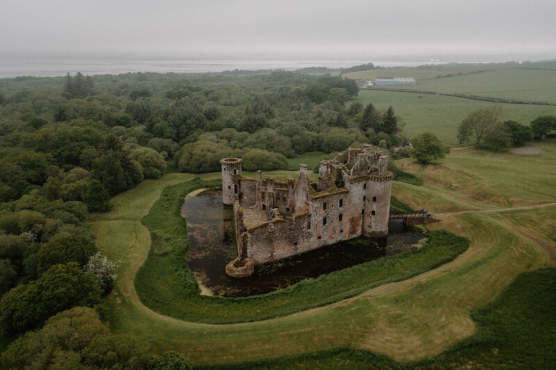 An aerial view shows the ruined red sandstone walls and towers of a moated castle surrounded by fields and woodland, its full moat visible.