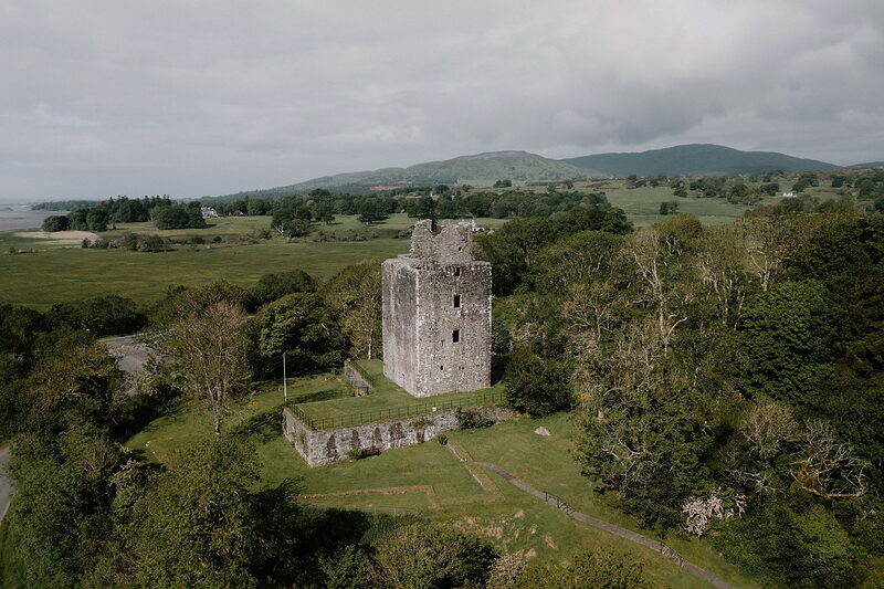 An aerial view shows a ruined stone tower keep standing in a field bordered by trees, hills and a river visible in the distance.