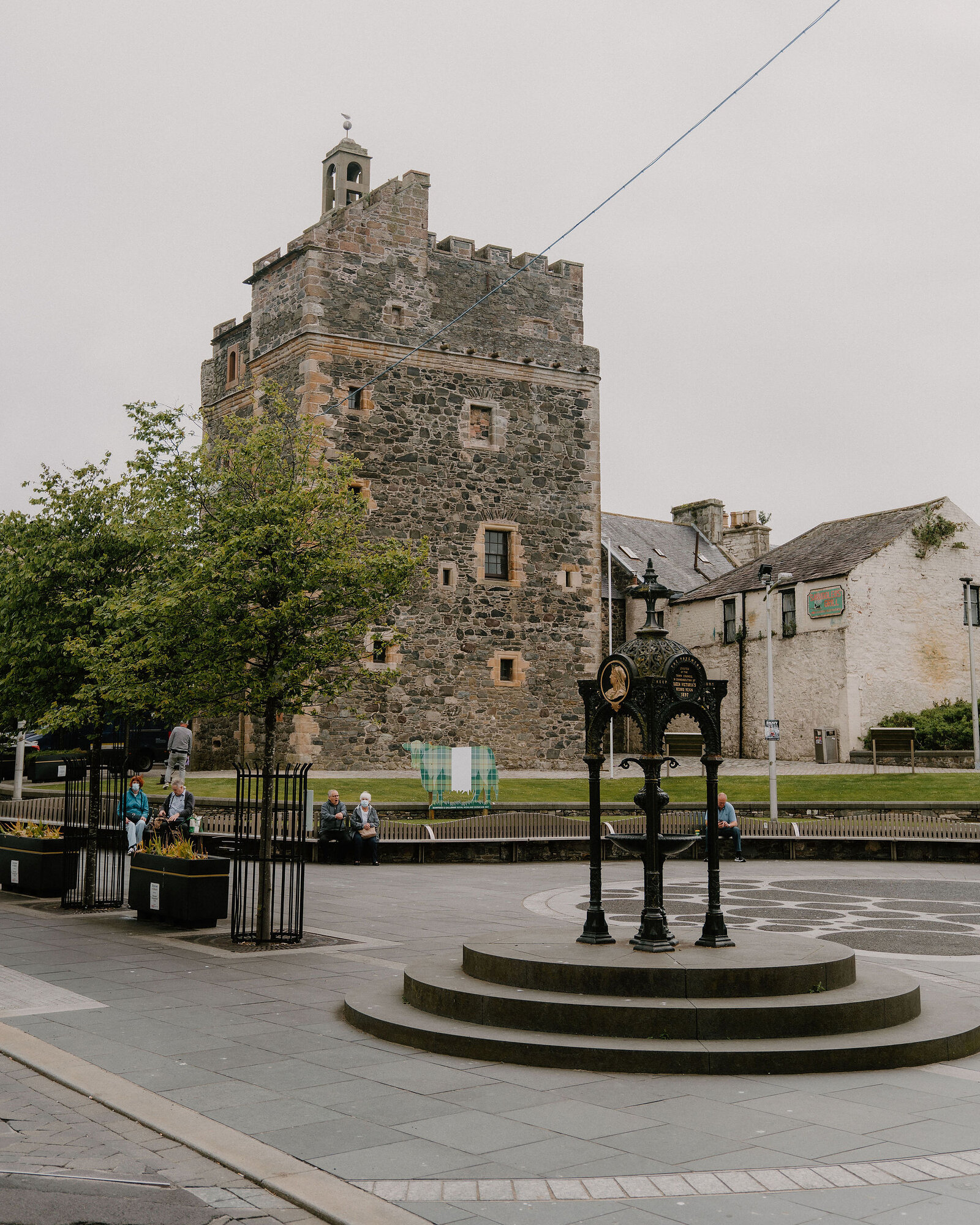 A stone tower keep with battlements rises behind a public drinking fountain in a paved square, people sitting on benches nearby.