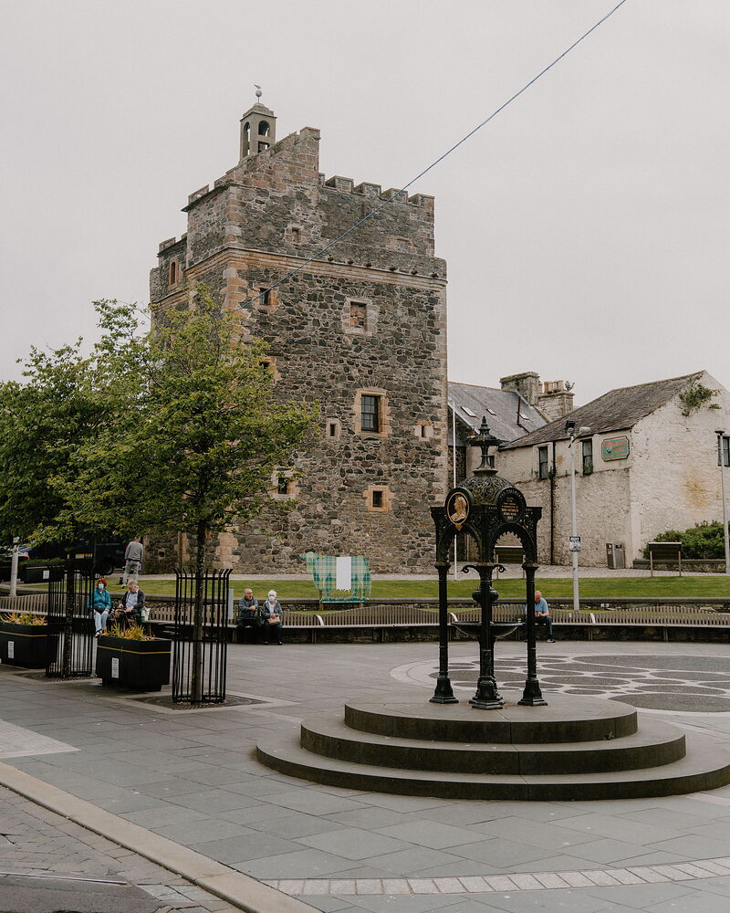 A stone tower keep with battlements rises behind a public drinking fountain in a paved square, people sitting on benches nearby.