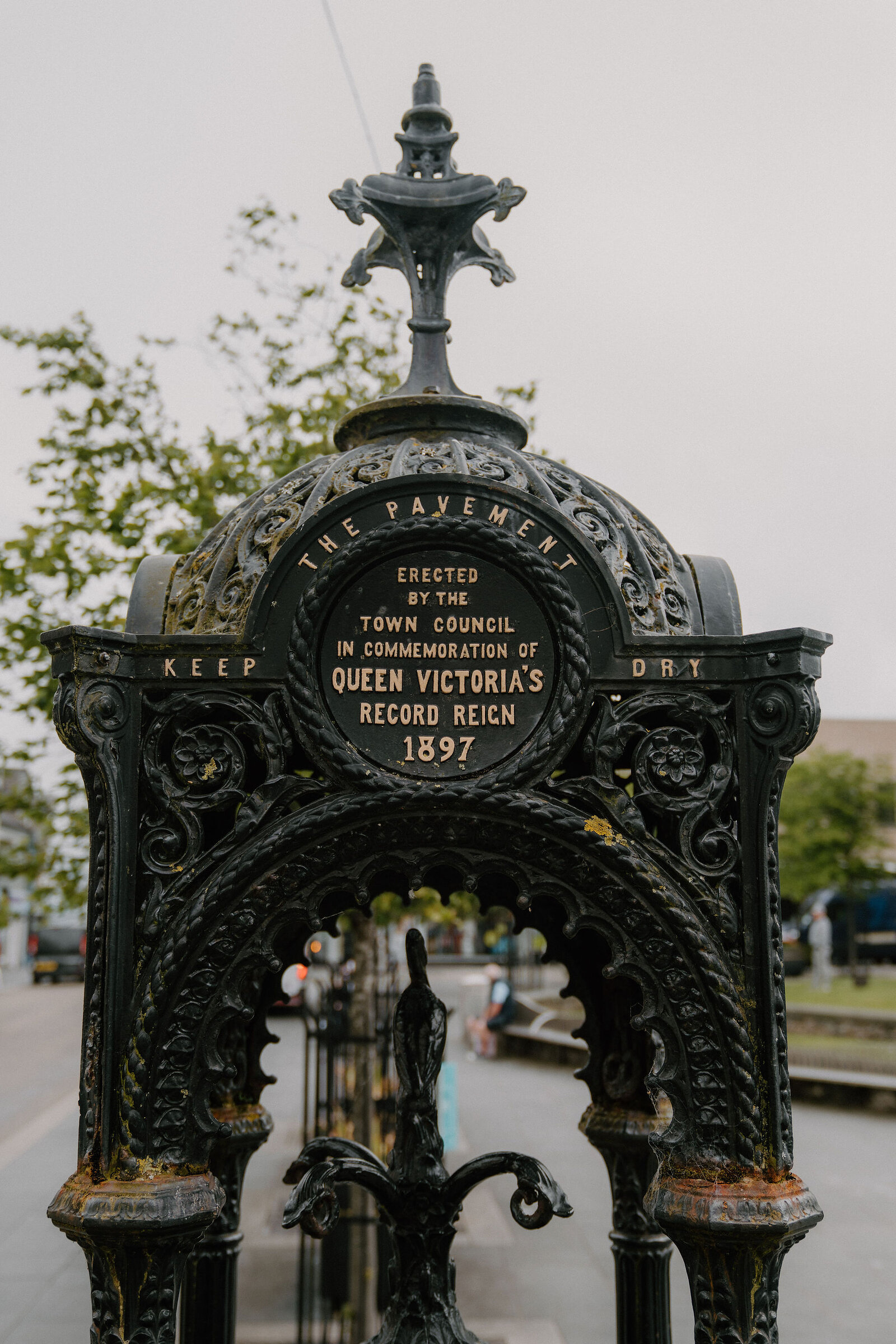 An ornate cast-iron drinking fountain inscribed "Erected by the town council in commemoration of Queen Victoria's record reign, 1897" stands in a paved square.