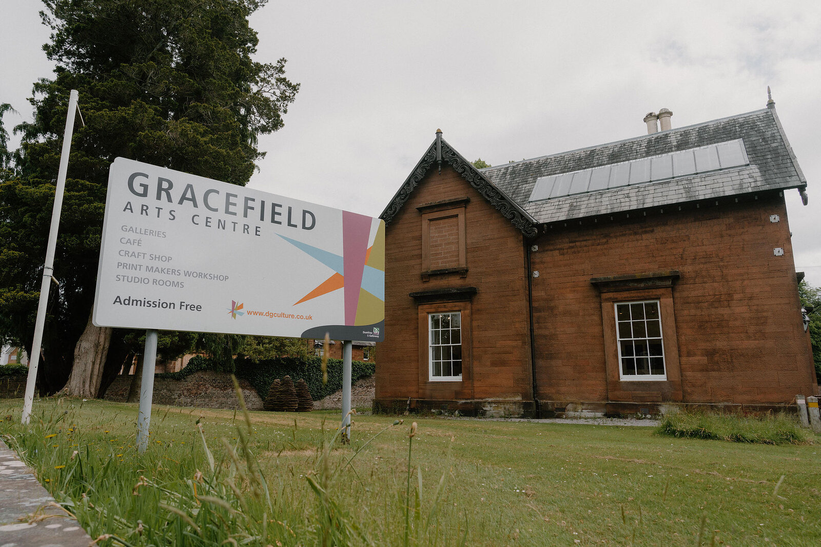A sign reading "Gracefield Arts Centre, gallery, cafe, craft shop, print makers workshop, studio rooms, admission free" stands beside a red sandstone building with solar panels on its roof.