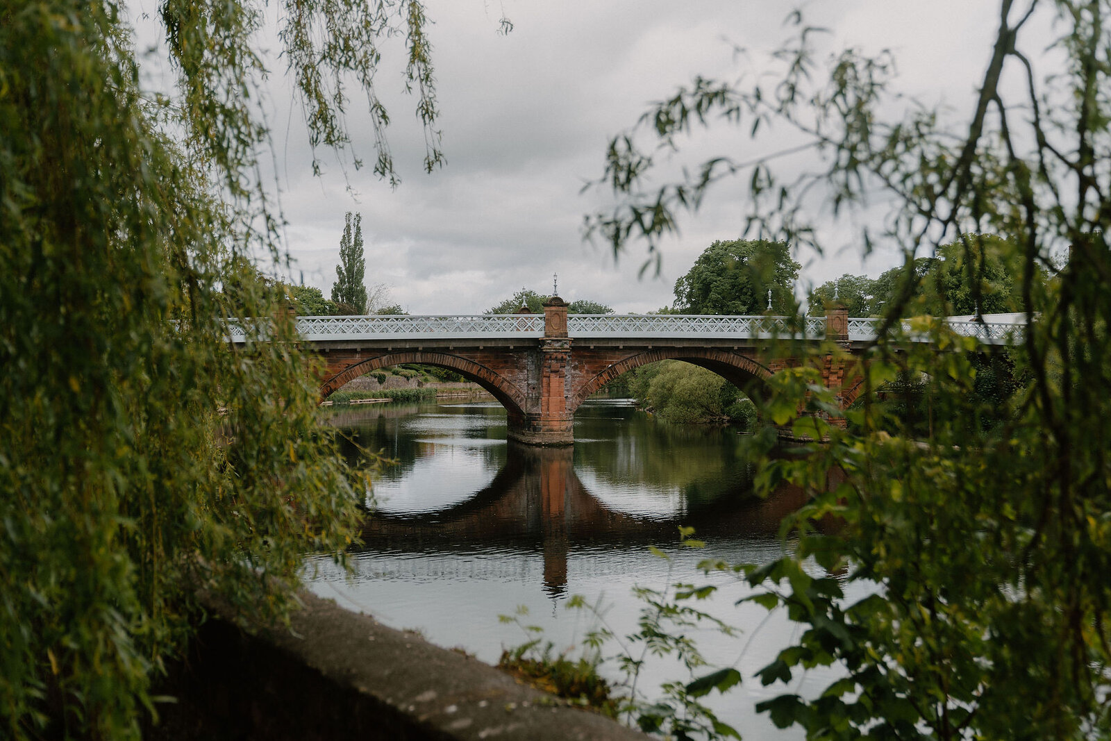 A metal truss bridge crosses a calm river, framed by overhanging willow trees on both banks.