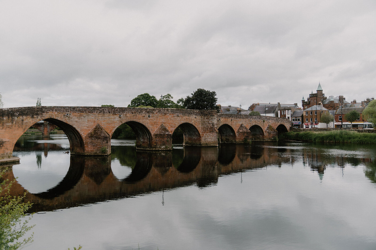 A multi-arched red sandstone bridge crosses a calm river, town buildings and a church spire rising along the far bank.