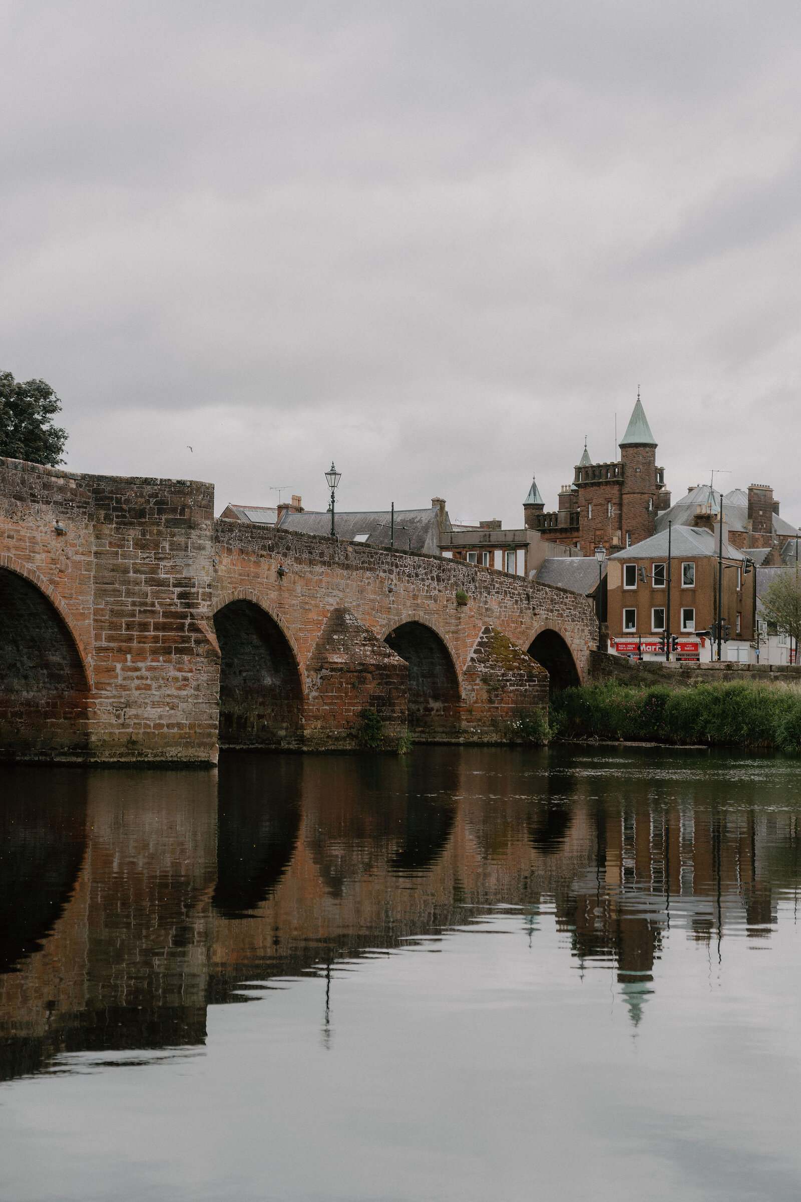 A multi-arched red sandstone bridge crosses a calm river, turreted town buildings reflected in the water behind.