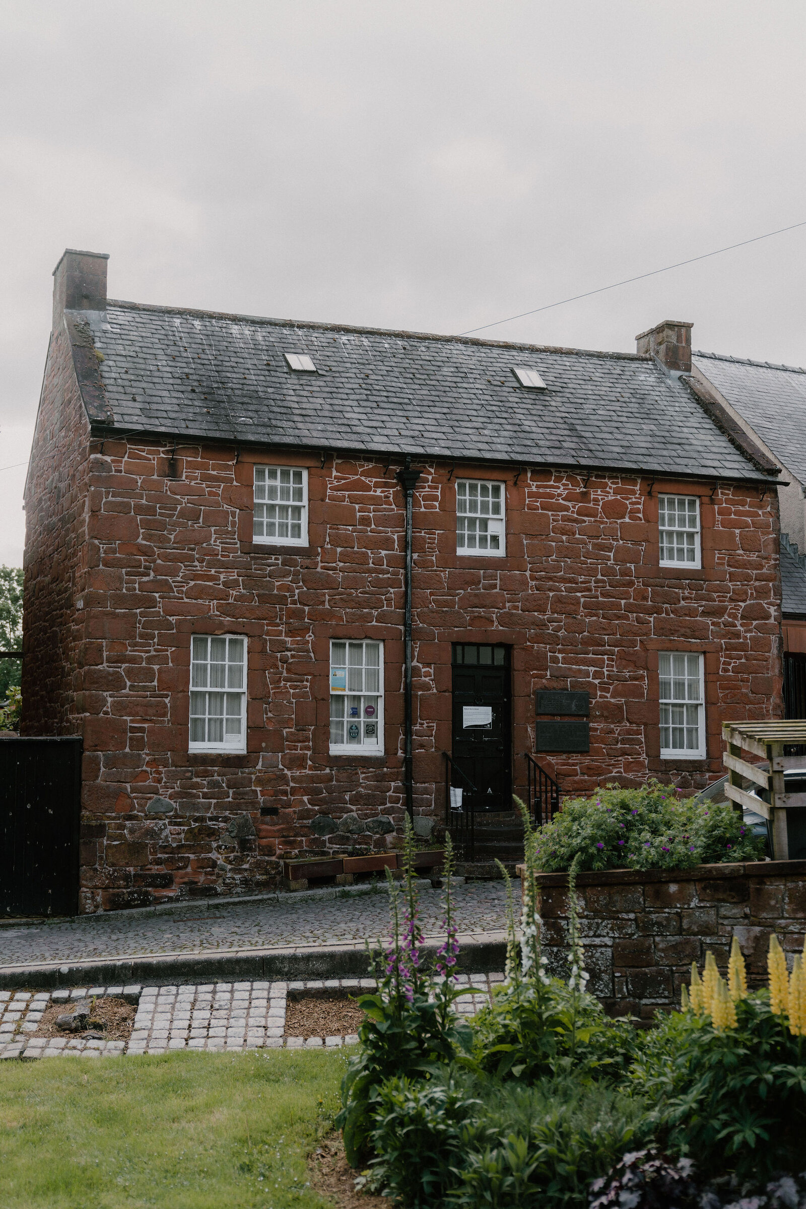 A red sandstone building with a slate roof stands behind a cobbled forecourt, foxgloves blooming in the garden bed in front.