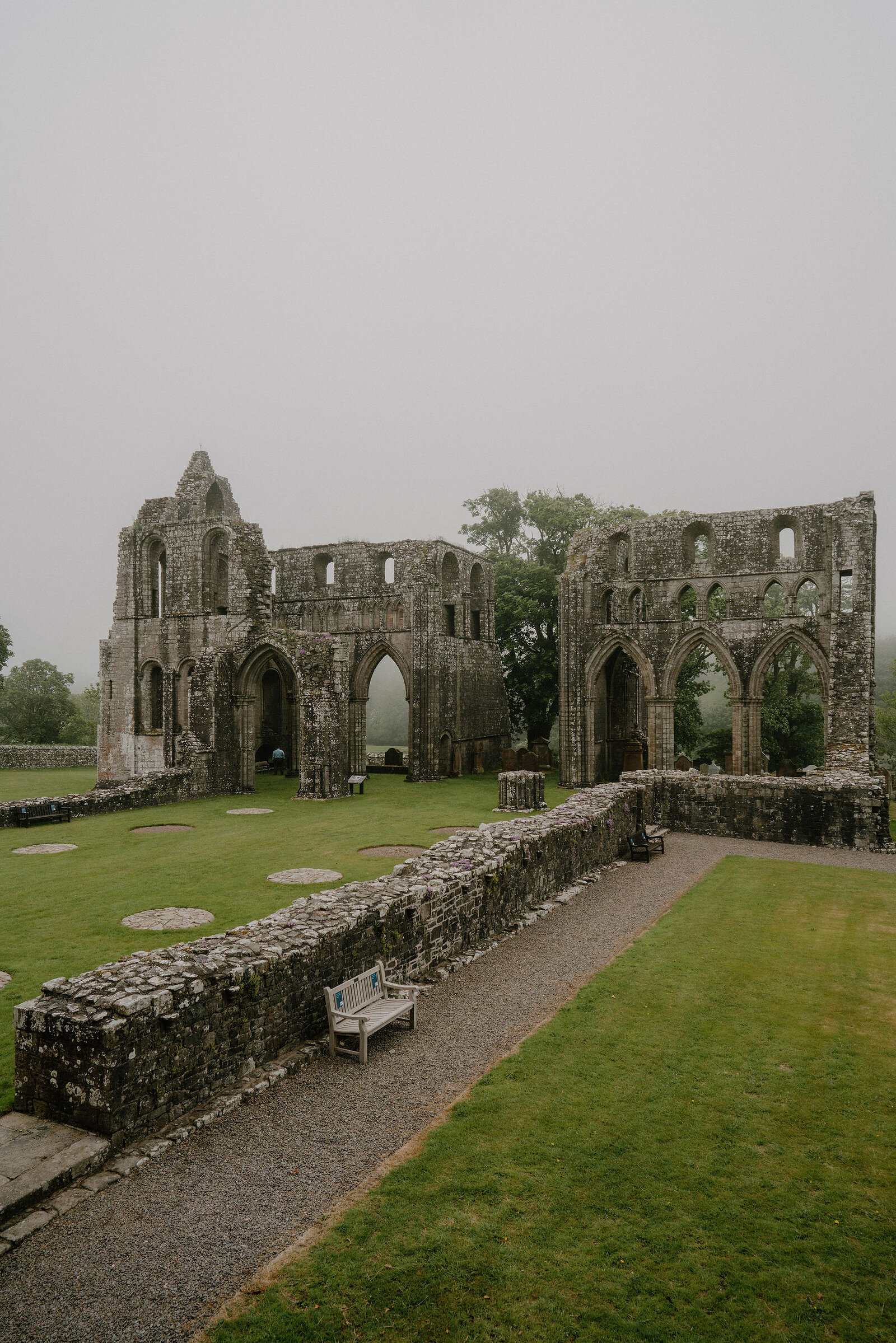 The ruined red sandstone walls and Gothic arches of an abbey stand in mist, a bench and gravel path lining the lawn in front.