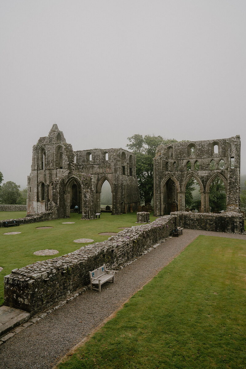 The ruined red sandstone walls and Gothic arches of an abbey stand in mist, a bench and gravel path lining the lawn in front.