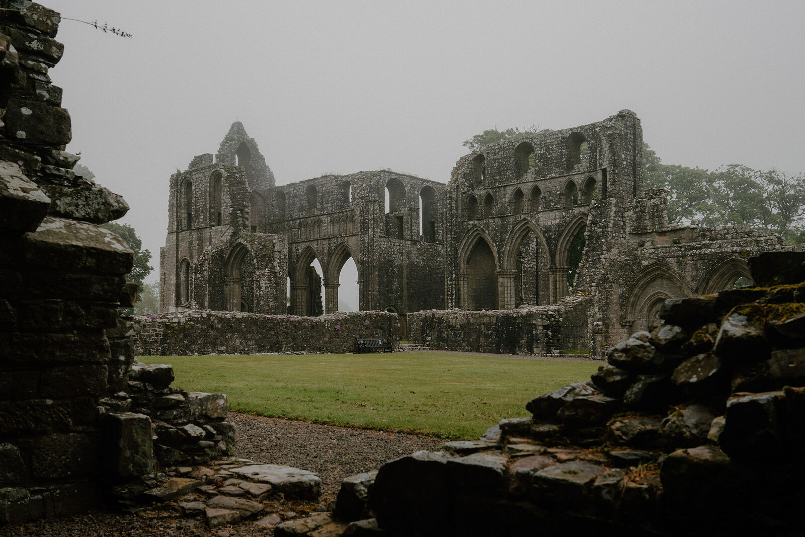 The ruined red sandstone walls and Gothic arches of an abbey stand in mist, seen through a stone gap in the foreground wall.