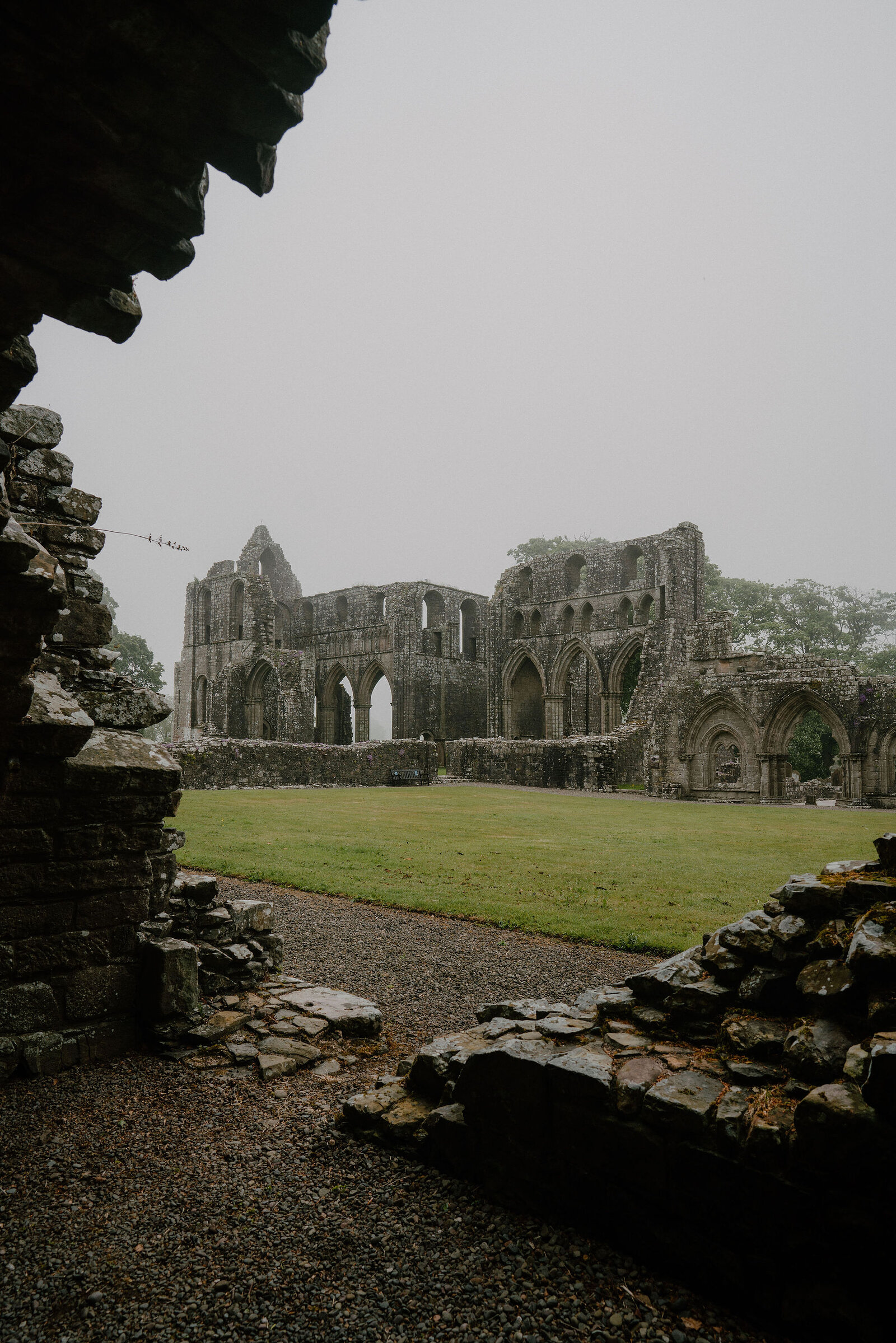 The roofless nave of a ruined abbey stands open to a grey sky, gothic arches and window tracery framed by a gravel path and mossy stone walls in the foreground, with cut grass filling the old floor.