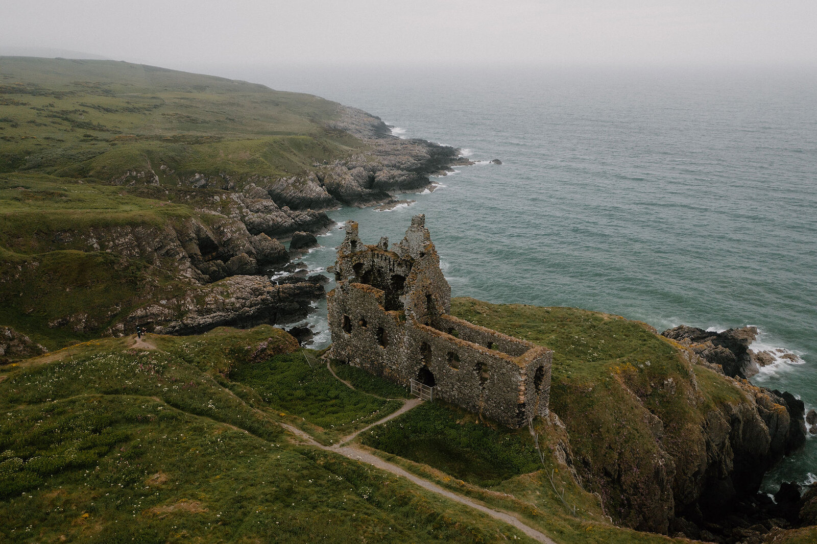 An aerial view along a green coastline shows a ruined castle standing on a rocky promontory, cliffs and coves stretching either side towards the sea.