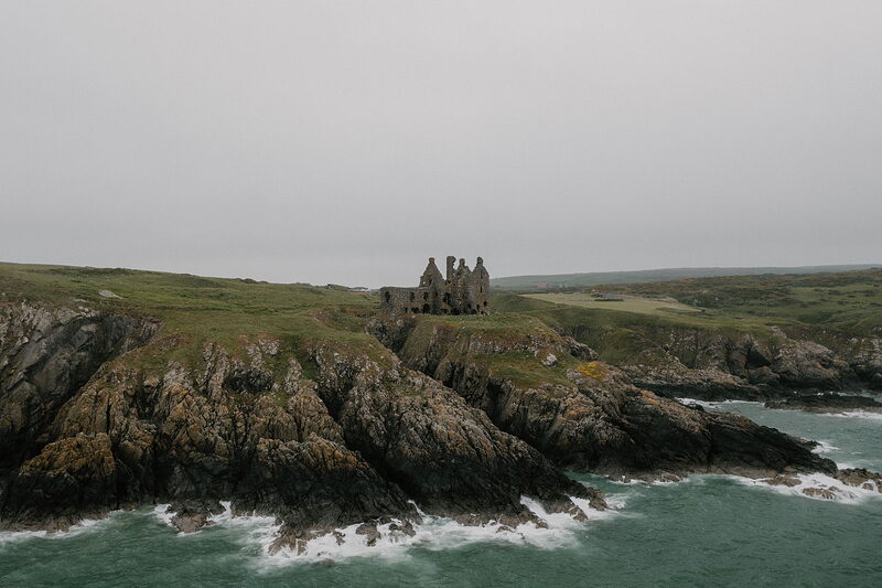 An aerial view shows a ruined castle standing on a rocky headland surrounded by turquoise sea, cliffs and grassy fields stretching along the coast.